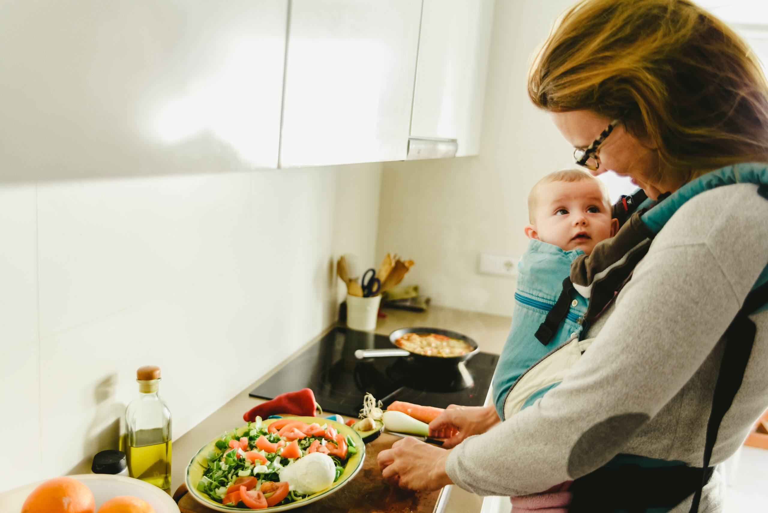 anya egy konyhában zöldségeket készít elő, a babája hordozóban előre van kötve, egymás szemébe néznek