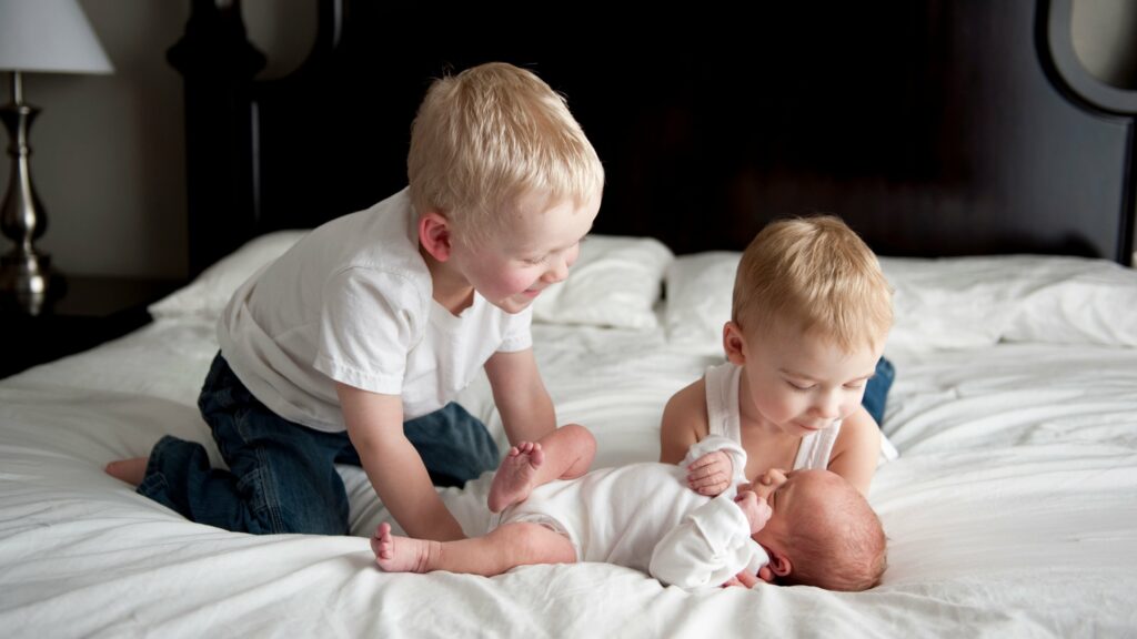 Két kisgyerek az ágyon, közöttük egy baba, akit megölelnek és mosolyognak rá